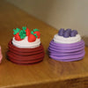 A stack of colorful fondant cakes, featuring red strawberries and purple frosting, displayed on a wooden surface.