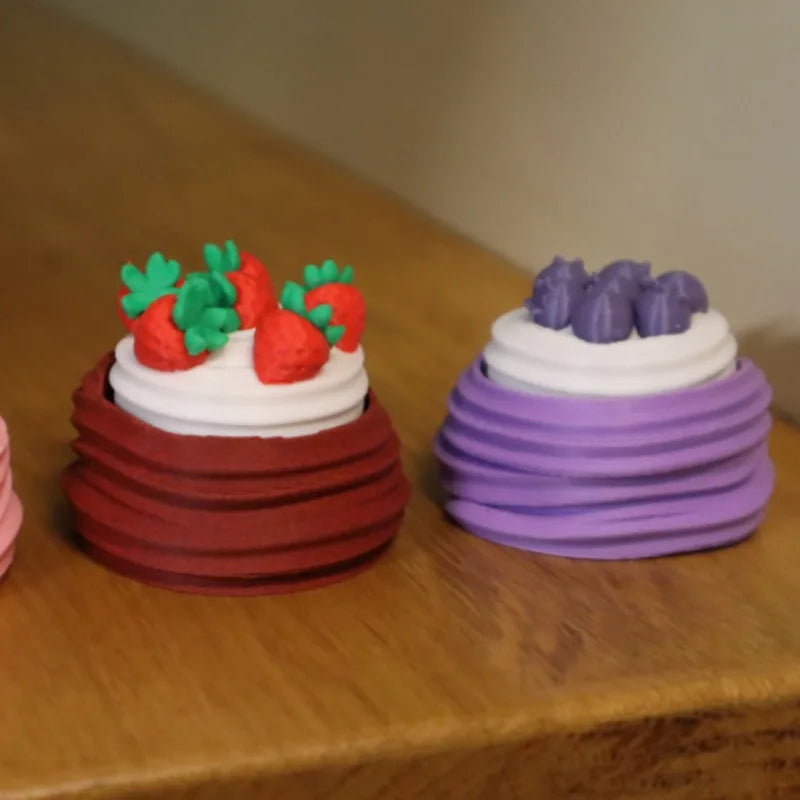 A stack of colorful fondant cakes, featuring red strawberries and purple frosting, displayed on a wooden surface.