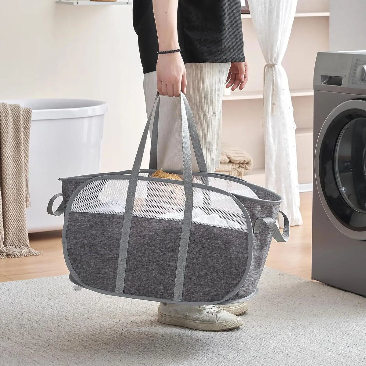 A gray collapsible laundry hamper with reinforced handles sits on a light rug next to a washing machine.