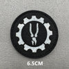 A black embroidered patch featuring a white gear design with a stylized hook and loop symbol.