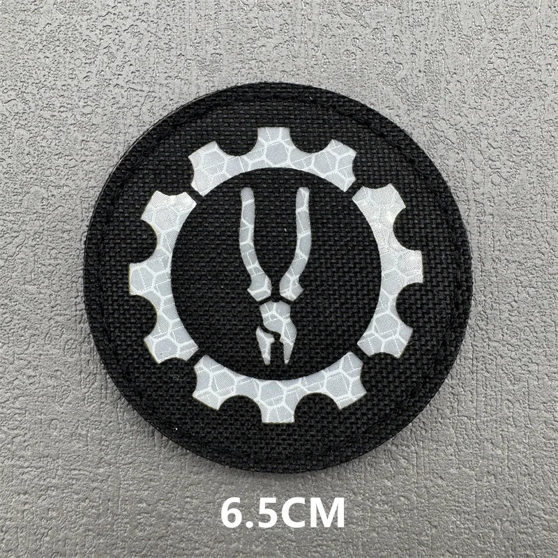 A black embroidered patch featuring a white gear design with a stylized hook and loop symbol.