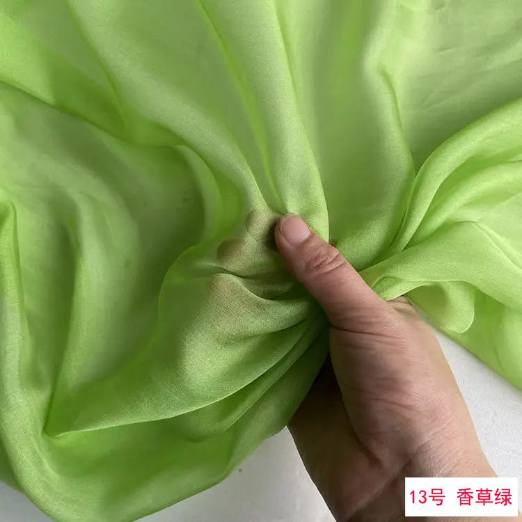 A hand is folding a piece of light green silk chiffon fabric to show its drape and texture.