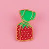 A glossy red strawberry enamel pin with a green fabric tie, displayed against a pink background.