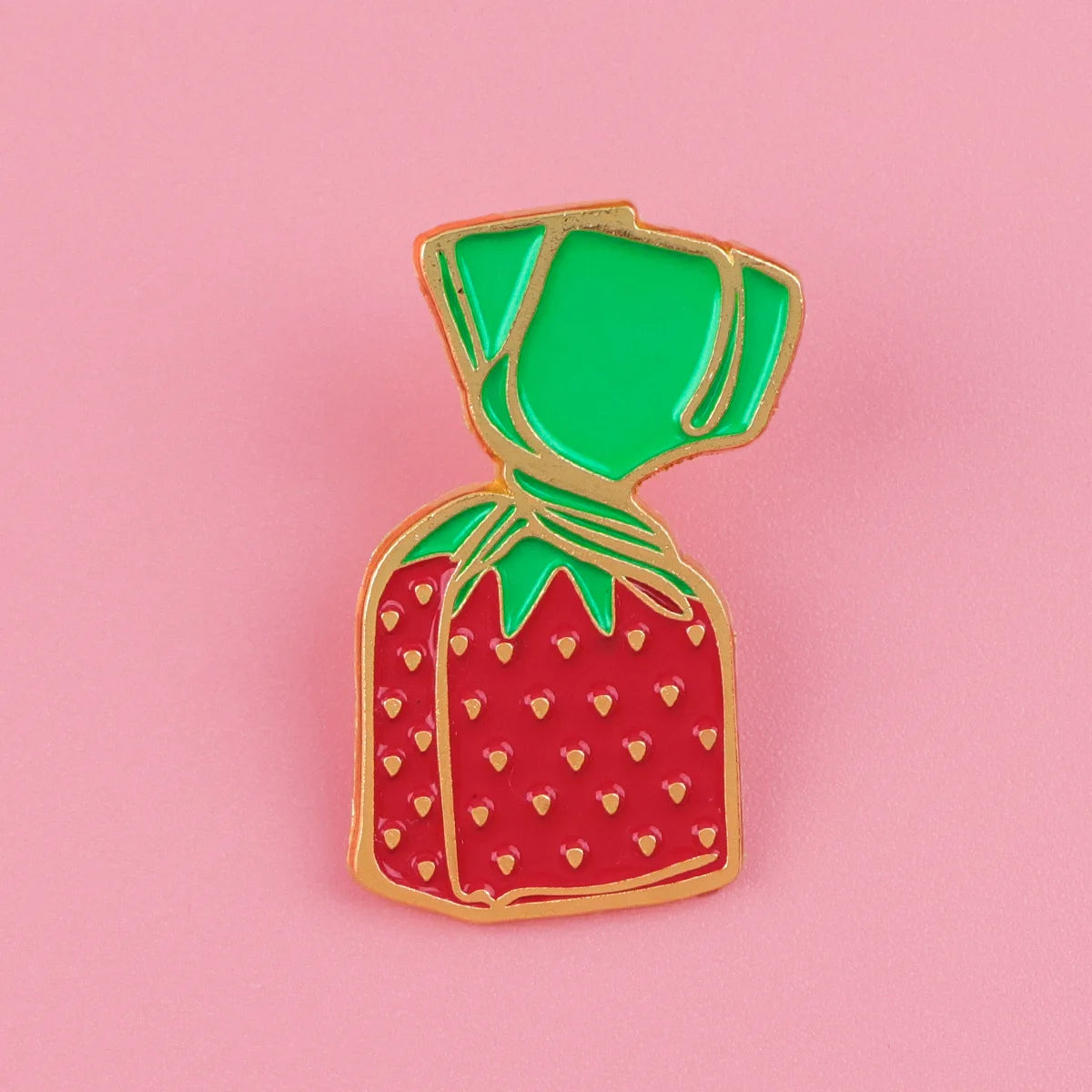 A glossy red strawberry enamel pin with a green fabric tie, displayed against a pink background.