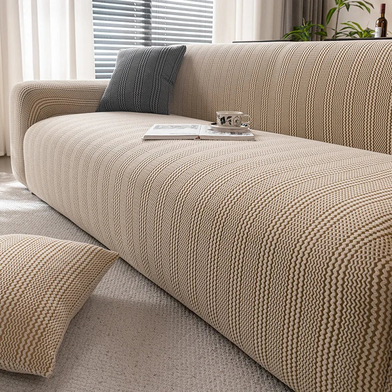 A beige quilted stretch sofa cover with a textured weave and a dark gray throw pillow on a light-colored sofa.