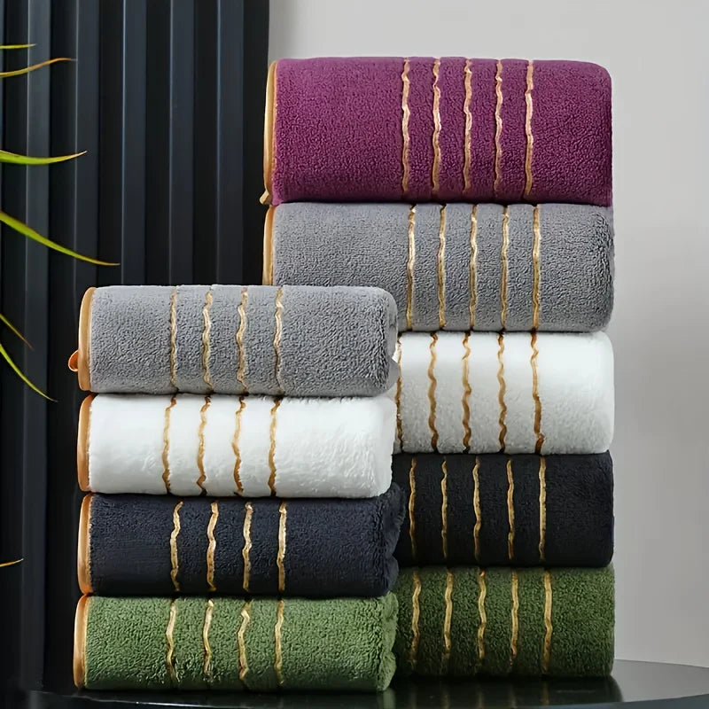 A stack of luxury bath towels in various colors including purple, gray, white, black, and olive green with gold stripes.