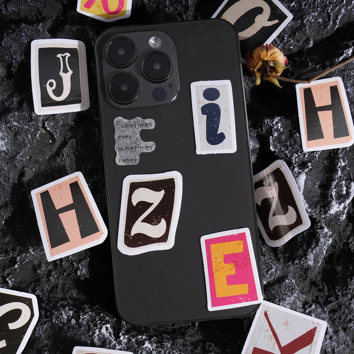A black smartphone is surrounded by various vintage alphabet stickers, including J, I, H, and Z.