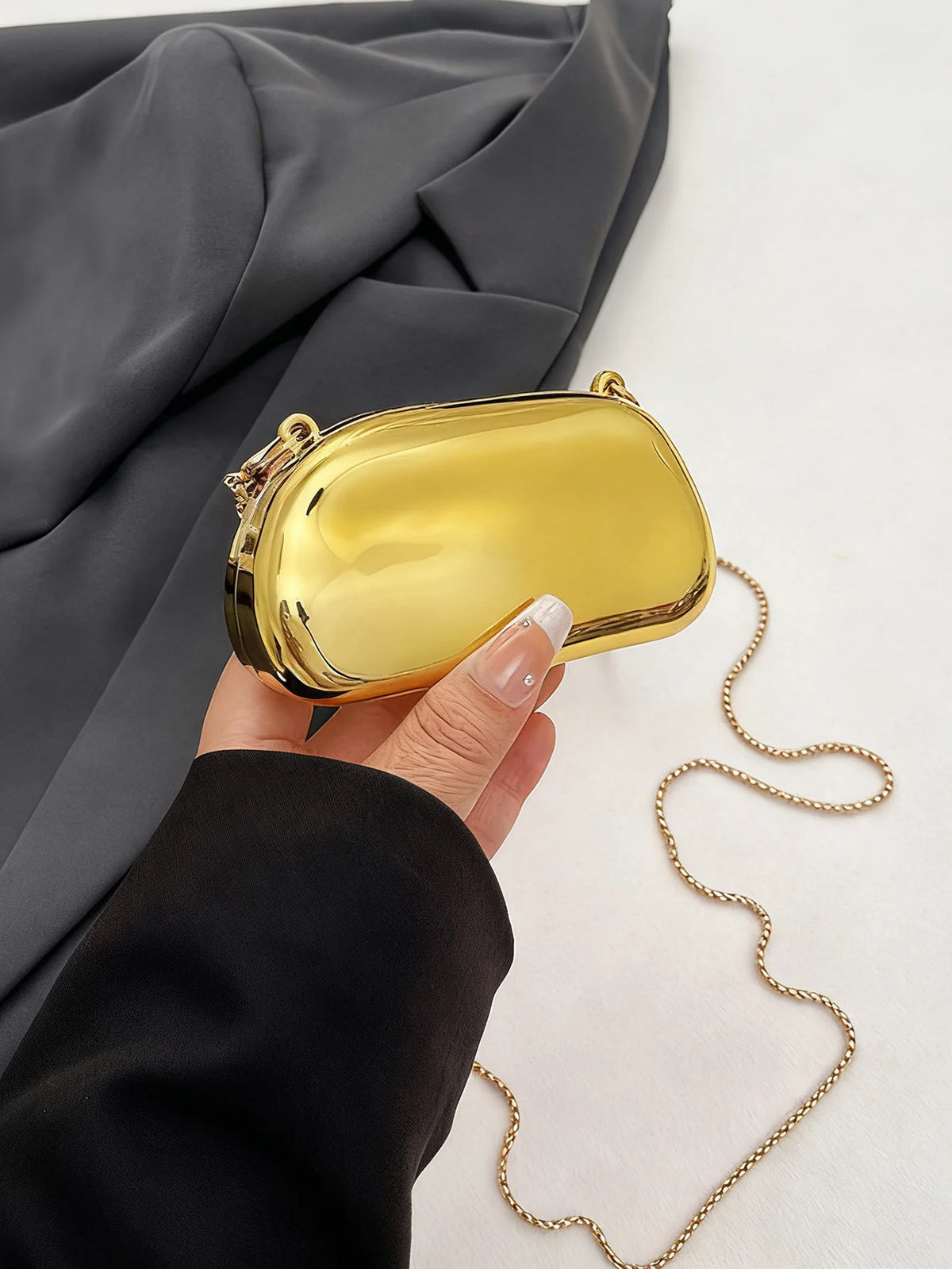 A hand holds a mini gold metal clutch bag against a dark gray fabric background.