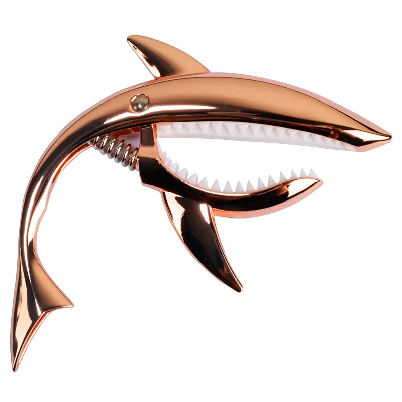 A bronze-colored guitar capo shaped like a shark, featuring a white plastic pick guard and serrated teeth.