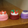 Three colorful cakes with frosting and fruit decorations are displayed on a wooden surface.