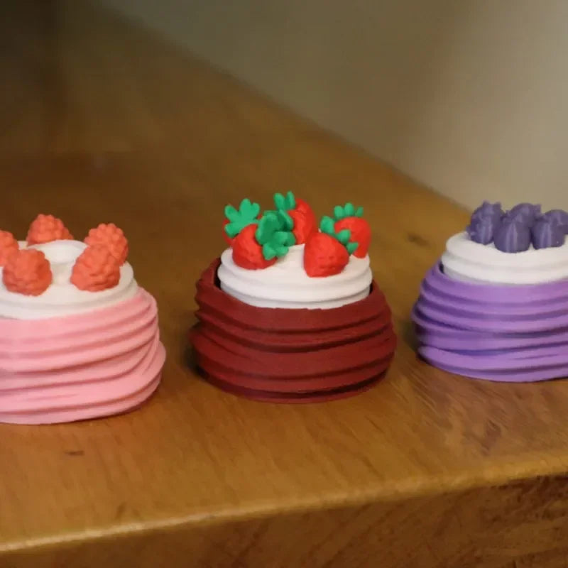 Three colorful cakes with frosting and fruit decorations are displayed on a wooden surface.