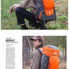 A woman wearing a tan jacket is shown sitting down with an orange and gray waterproof hiking backpack in a grassy outdoor set