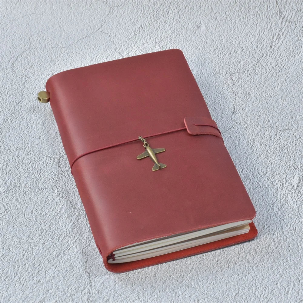 A closed, reddish-brown leather journal with a brass airplane-shaped clasp resting on a white textured surface.
