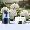 A black smart car diffuser with ambient light and auto mist sits on a white surface next to white roses.