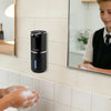 A boy is using a black touchless automatic soap dispenser on a bathroom counter in a school setting.