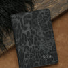 Black leopard print passport holder with personalized name embossing resting on a brown surface.
