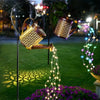 A vintage solar watering can garden light with a shepherd's hook illuminates a cascade of glowing fairy lights over purple fl