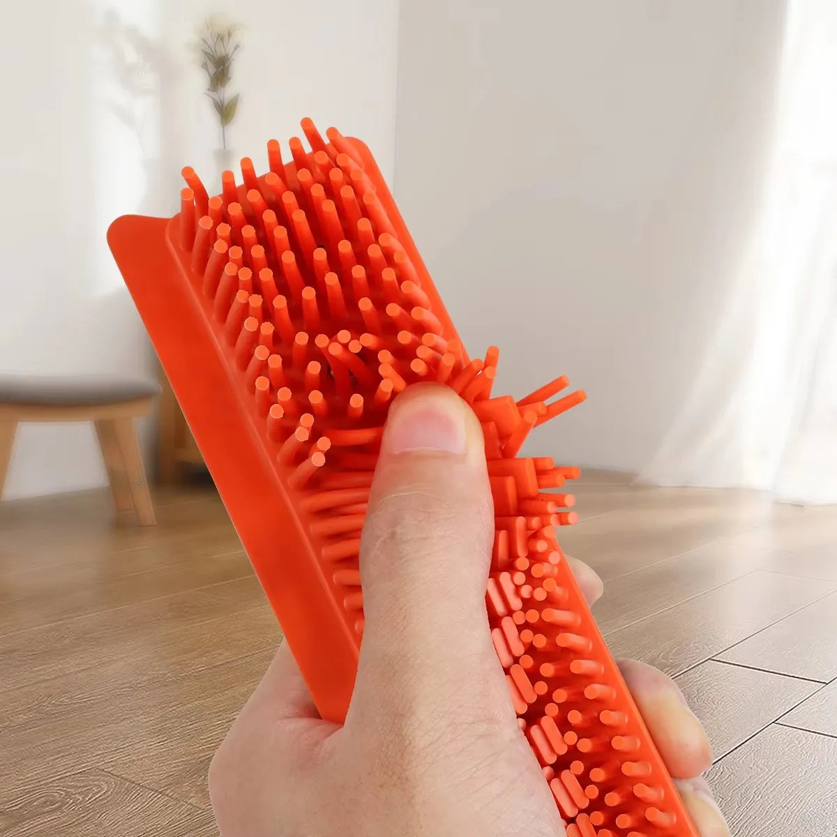 A hand holds an orange rubber broom and carpet rake with numerous bristles against a light background.
