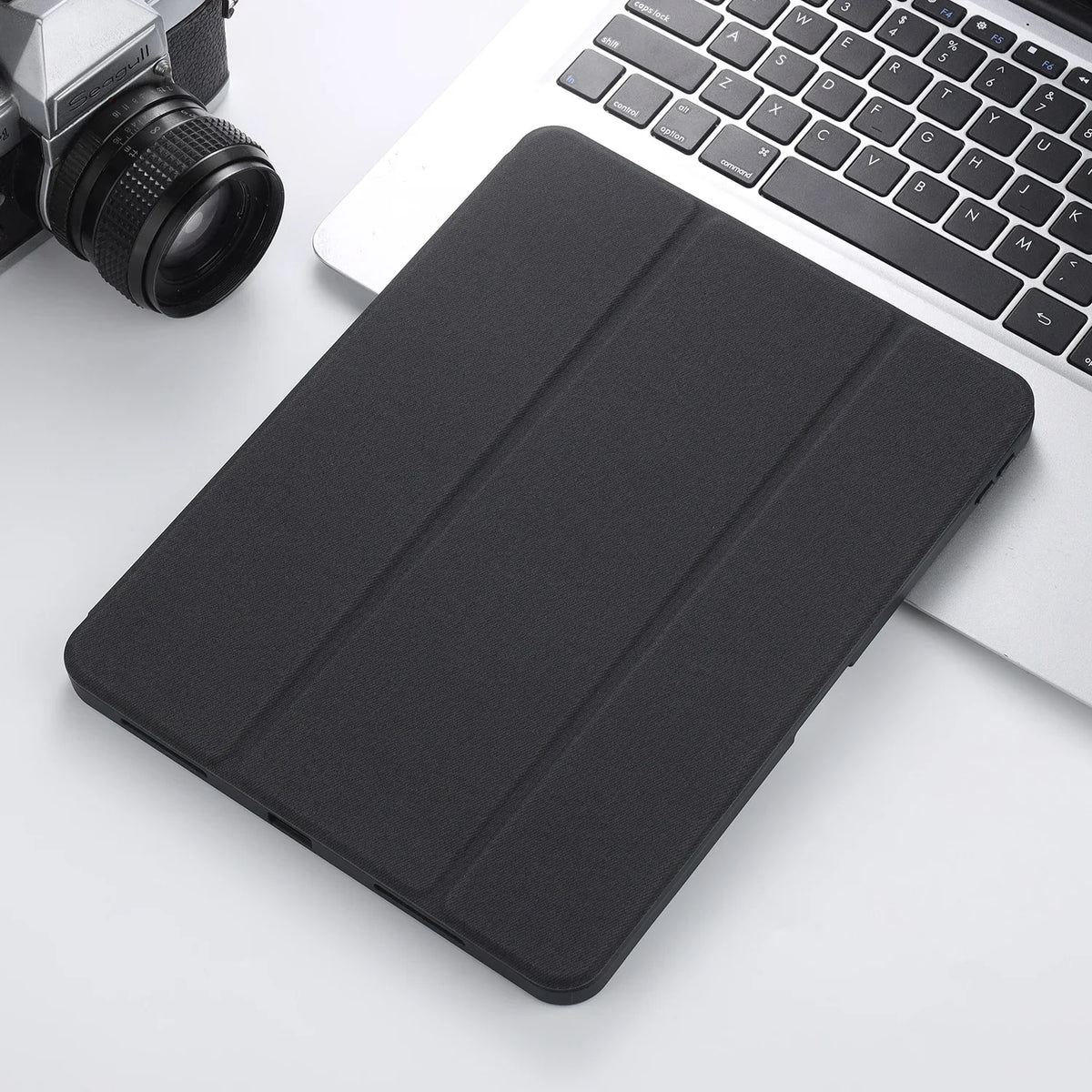 A black Retro Marble iPad Pro 12.9 case with a pencil holder resting on a white surface next to a silver camera and a laptop.