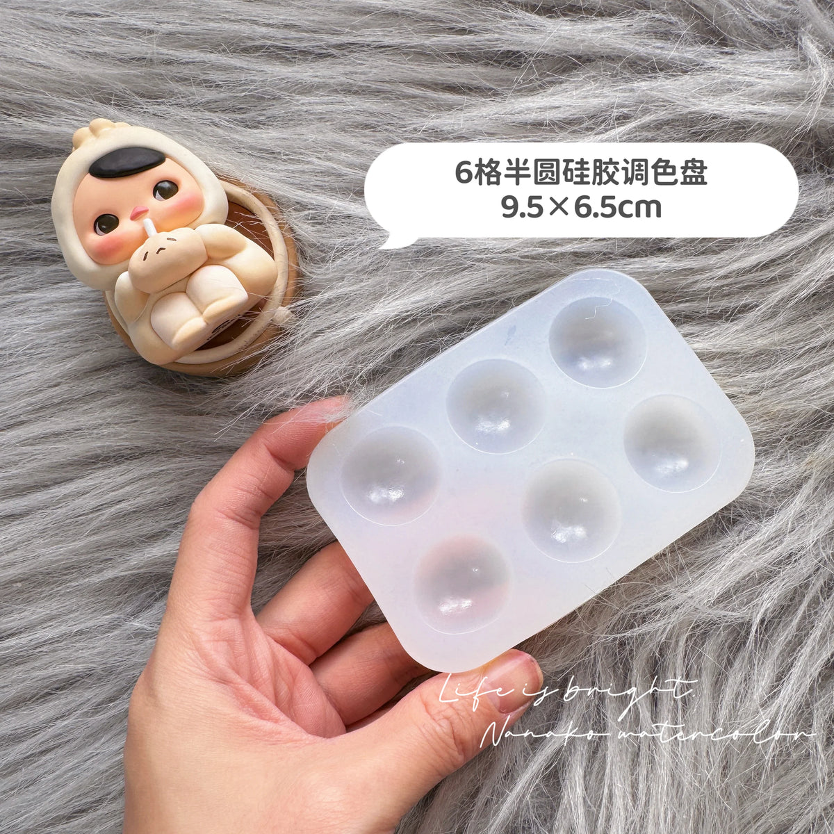 A hand holds a silicone paint palette tray with six wells, next to a small, round silicone figure.