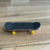 A black wooden fingerboard skateboard with yellow trucks resting on a light wooden surface.