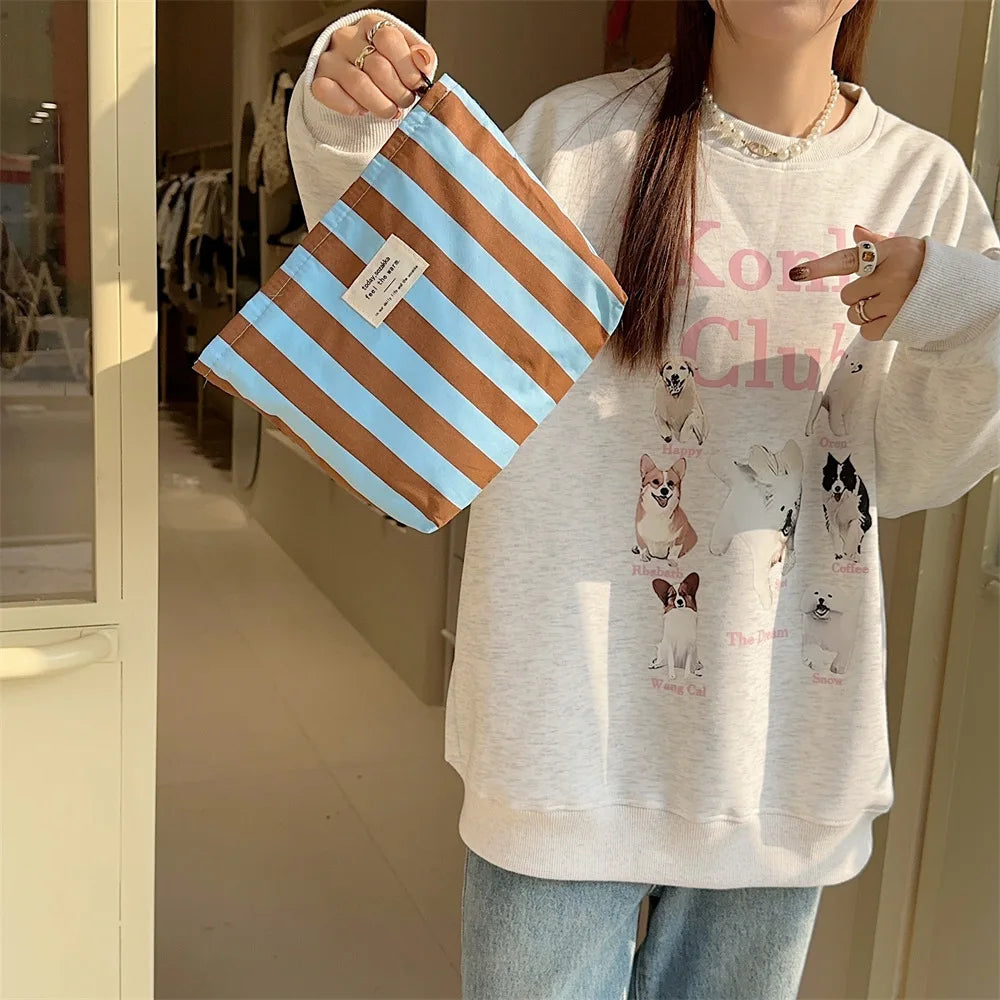 A person holds a striped canvas cosmetic bag with large capacity in front of a light gray sweatshirt.