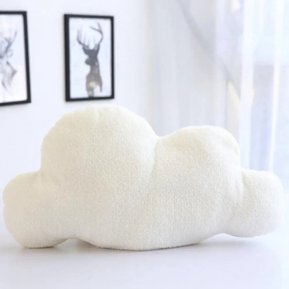 A fluffy white cloud-shaped plush throw pillow resting on a white surface next to framed artwork.