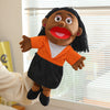 A hand puppet with dark skin, black hair, and orange clothing is being held by a person in a light-colored sleeve.