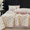 A light beige floral quilted bedspread with a pink trim is draped over a dark bed, next to a white pillow.