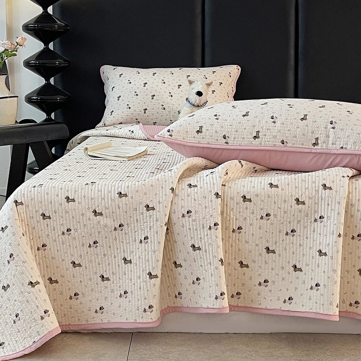 A light beige floral quilted bedspread with a pink trim is draped over a dark bed, next to a white pillow.