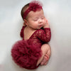 A newborn baby wearing a deep red lace princess tutu romper and headband, lying down.