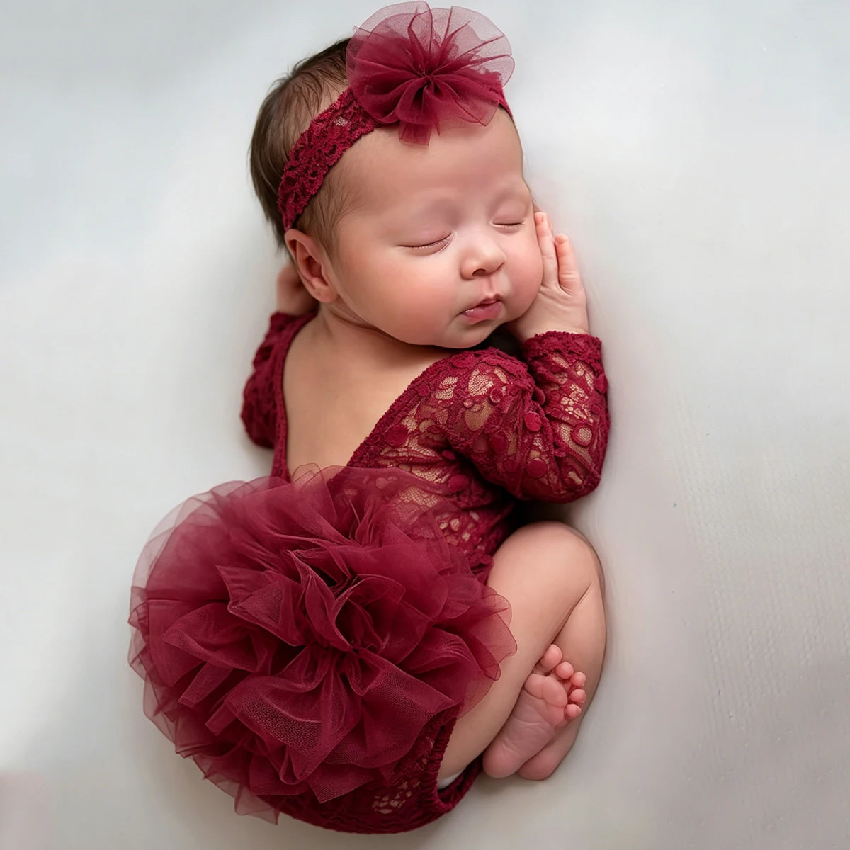 A newborn baby wearing a deep red lace princess tutu romper and headband, lying down.
