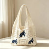 A cream-colored handwoven knit tote bag with a white crochet pattern and blue bear embroidery resting on a light surface.