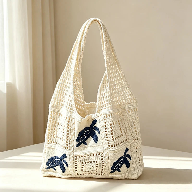 A cream-colored handwoven knit tote bag with a white crochet pattern and blue bear embroidery resting on a light surface.