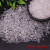 A pile of clear quartz crystal chips scattered on a dark surface with a green leaf in the background.