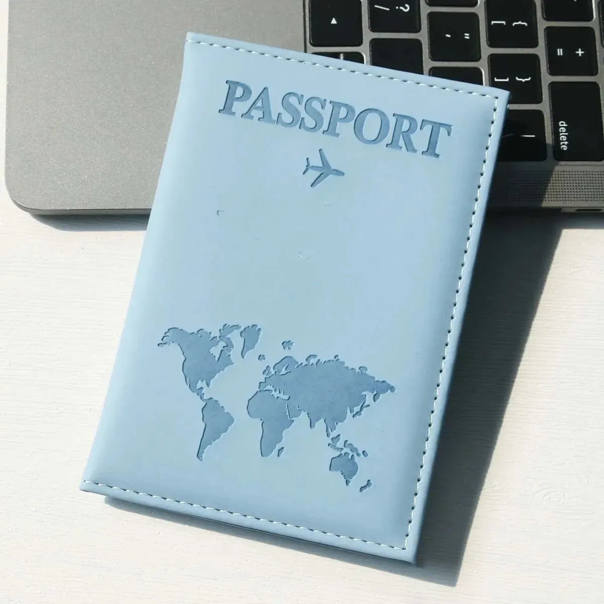 Hellblaue Leder-Reisepasshülle mit Weltkarten-Design neben silbernem Laptop und schwarzer Tastatur