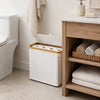 A white bamboo-framed toilet paper storage basket with a lid sits next to a toilet and on a wooden shelf.