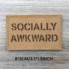 A rectangular tan woven patch with the text SOCIALLY AWKWARD printed in black capital letters on a wooden surface.