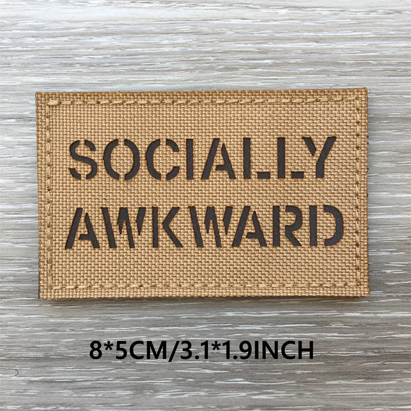 A rectangular tan woven patch with the text SOCIALLY AWKWARD printed in black capital letters on a wooden surface.