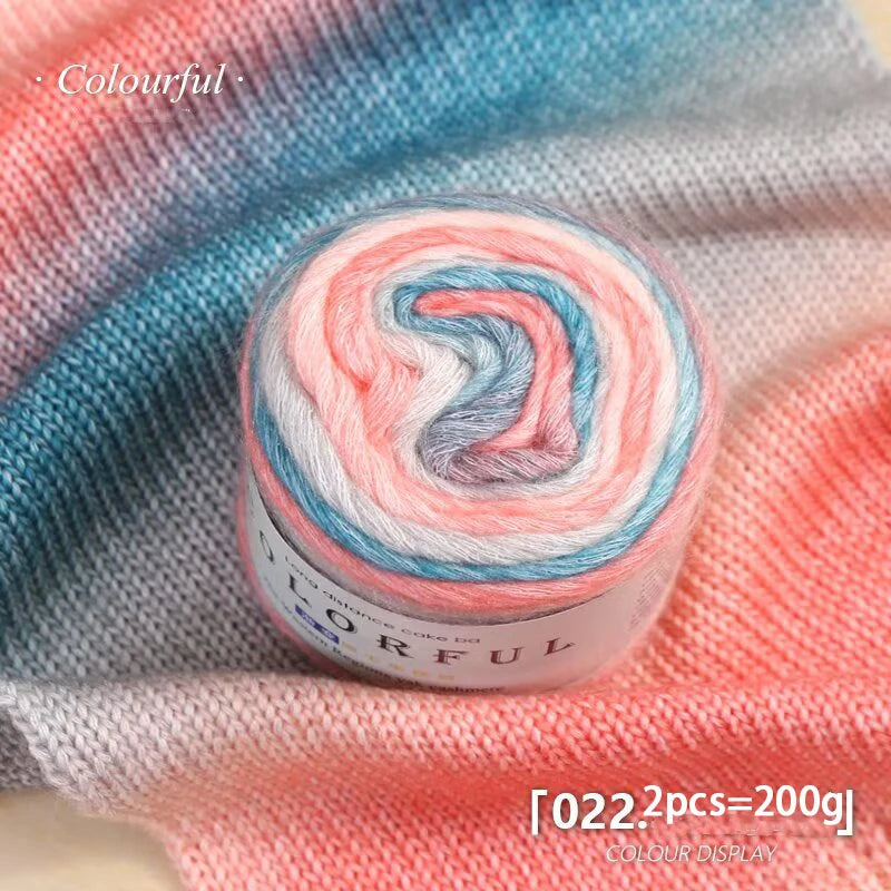A spool of colorful gradient cake yarn showing pink, blue, and white strands against a light fabric background.