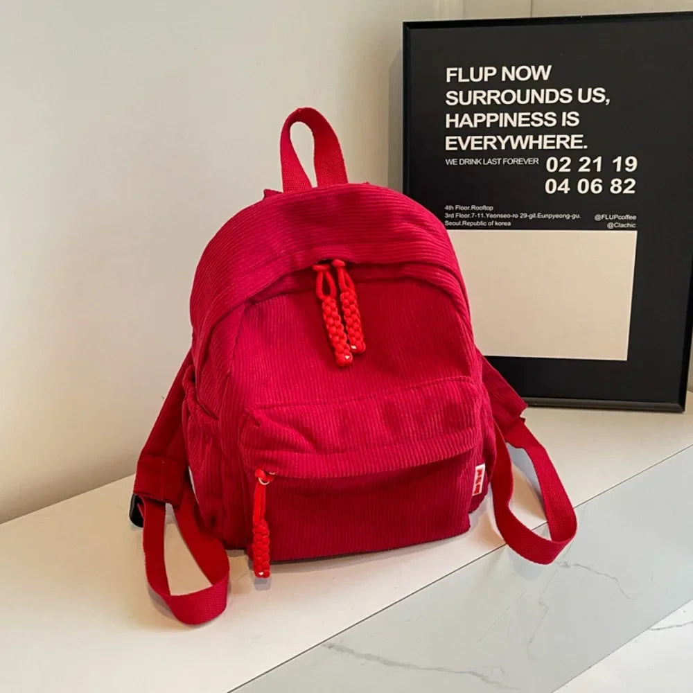 A bright red corduroy backpack is displayed on a white surface next to a framed sign.