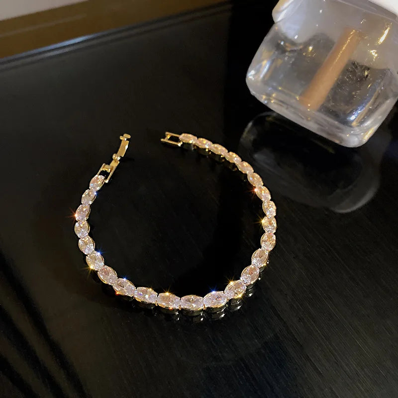 Nahaufnahme eines zarten goldenen Tennisarmbandes in einer Glasverpackung vor dunklem Hintergrund.