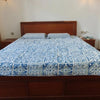 A wooden bed frame with a blue and white patchwork cotton bedspread is shown in a bright bedroom.