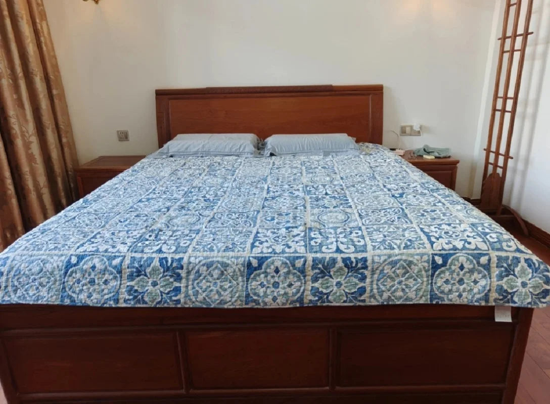 A wooden bed frame with a blue and white patchwork cotton bedspread is shown in a bright bedroom.