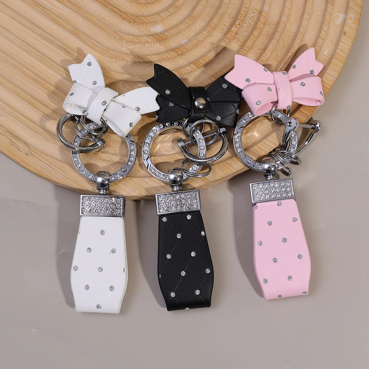 Three PU leather bow keychains in white, black, and pink with O-shaped buckles displayed on a wooden surface.