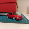 A red 1:64 scale alloy car model is displayed on a grid surface next to a red box.