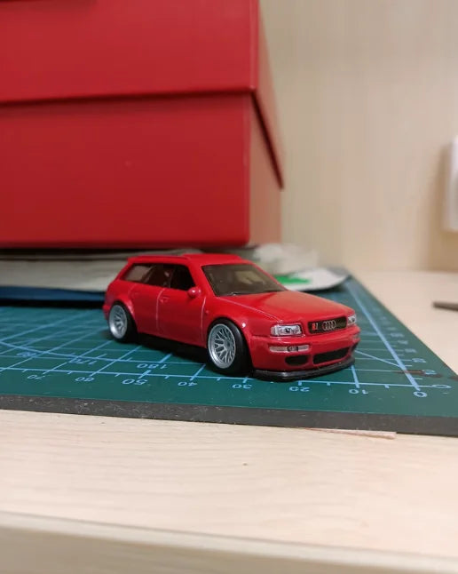 A red 1:64 scale alloy car model is displayed on a grid surface next to a red box.