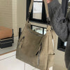 A hand holds a beige matte leather crossbody hobo bag with an adjustable strap against a white and gray background.