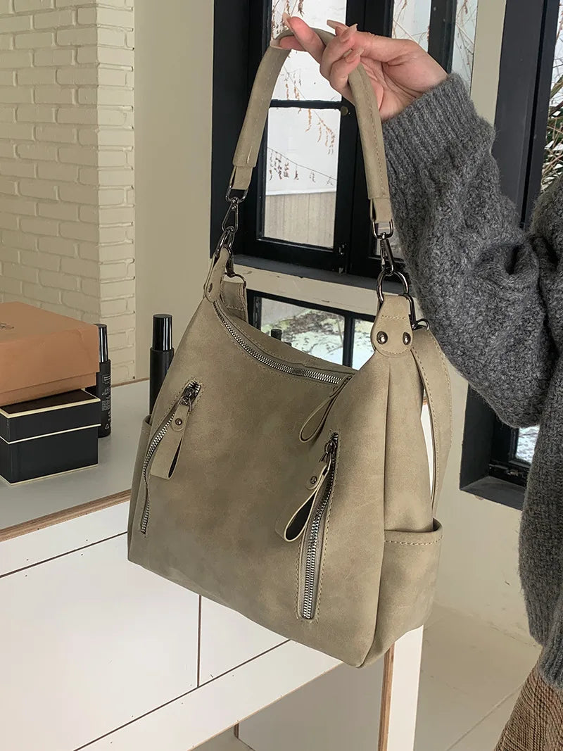 A hand holds a beige matte leather crossbody hobo bag with an adjustable strap against a white and gray background.