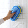 A hand is holding a blue and yellow sponge cleaning brush against a white tiled wall.